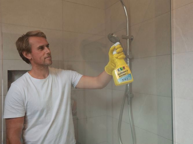 A gentleman holding a bottle of CLR Bathroom & Kitchen Foaming Action Cleaner next to a shower screen in a bathroom.