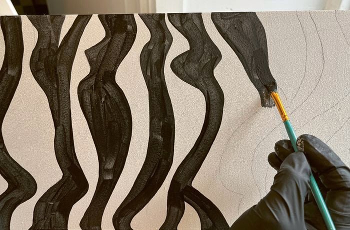 A gloved hand paints black zebra stripes onto a white surface with a paintbrush.