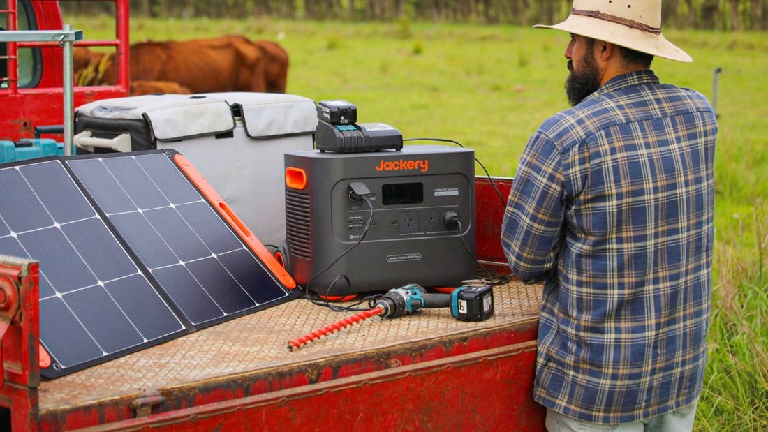 : The back of a male wearing a wide-brimmed hat and a check shirt standing in front of a Jackery portable power station on the back of a ute in a farm setting.