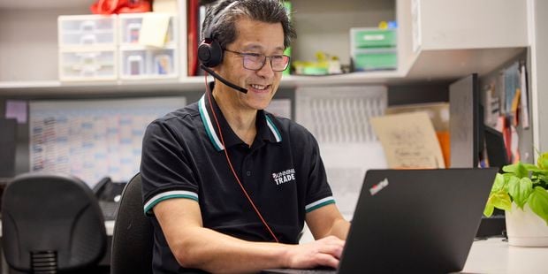 Bunning Trade team member wearing a headset types on a laptop at a desk with shelves of folders and supplies in the background.