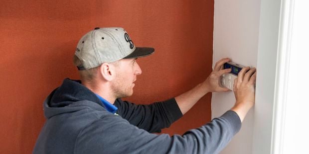 a person wearing a cap and hoodie is using a level to install an electrical outlet on a white wall