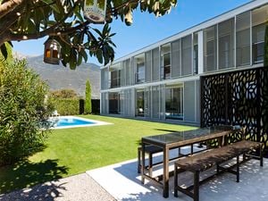 "Modern outdoor patio with a glass-top table and benches in front of a two-story building with large windows and sliding shutters, next to a lawn and in-ground pool