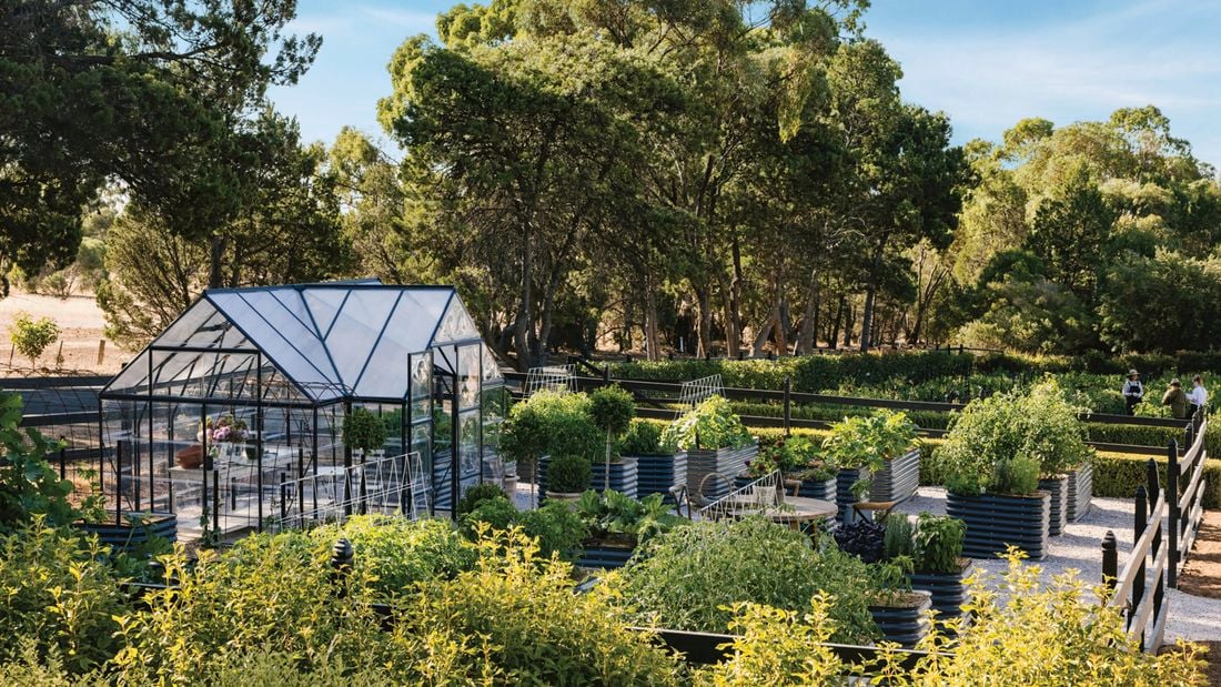 A greenhouse in a lush garden with raised vegetable beds and trees.