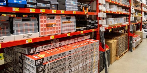 Shelving unit in Bunnings displaying storage and organisation products, including plastic parts bins, compartment organisers, toolboxes and boxed adjustable shelving. Products are arranged on red industrial racks with price labels along each shelf.