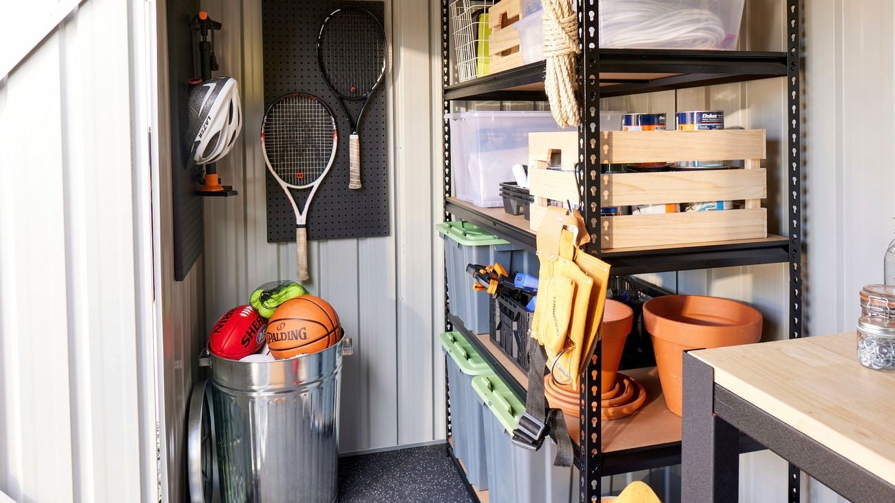 Inside of a garden shed with wooden shelves holding storage containers and tennis rackets hanging on a pegboard