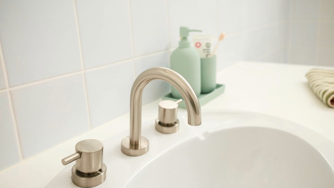 Close-up of a white bathroom sink with a curved brushed metal faucet and matching taps, set into a white benchtop, with green-toned soap dispensers and toothbrush holder arranged on a tray in the background against white tiled walls.