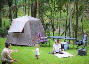 A camping setup in the bush with a tent, chairs, a table, a Jackery power station and a solar panel. A man kneels reaching towards a toddler, while a woman sits on a picnic mat.