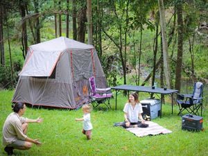 A camping setup in the bush with a tent, chairs, a table, a Jackery power station and a solar panel. A man kneels reaching towards a toddler, while a woman sits on a picnic mat.