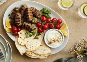 Lamb kofta on a white platter with pita bread, a bowl of creamy garlic sauce, cherry truss tomatoes and lemon wedges garnished with oregano.