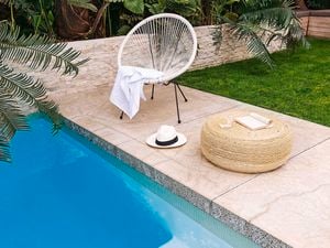 Outdoor lounge chair, hat and book next to edge of swimming pool in backyard with tropical plants