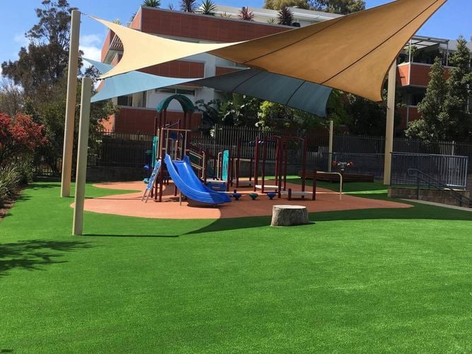 Outdoor playground with artificial grass, wooden swing set with blue net swing, slide, green shade canopies, and surrounding trees and fence.