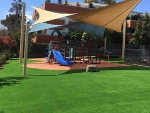 Outdoor playground with artificial grass, wooden swing set with blue net swing, slide, green shade canopies, and surrounding trees and fence.