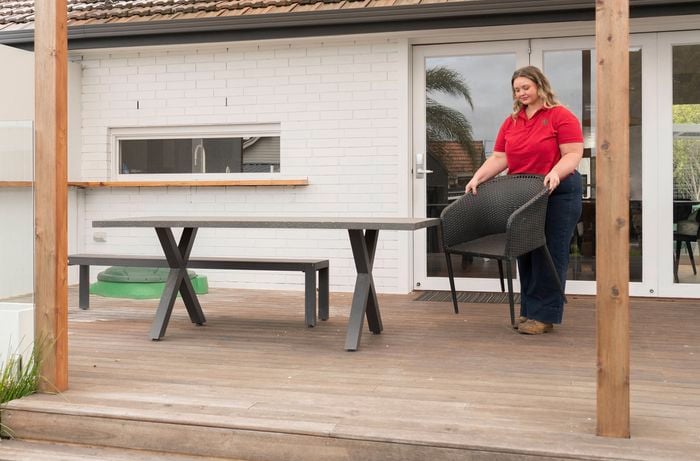 A Bunnings Team Member is standing on a wooden deck, holding a black chair and arranging it near a table