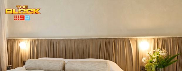 A furnished bedroom with a bed, side table with a vase on it and a bedhead behind it. And The Block logo in the image.