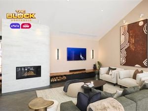 A modern living room with a high ceiling, featuring a fireplace on the left with firewood stored beneath. The space includes a green sofa, white armchairs, and a small round coffee table with books. Two large pieces of wall art are displayed—one above the fireplace and another on the right wall.