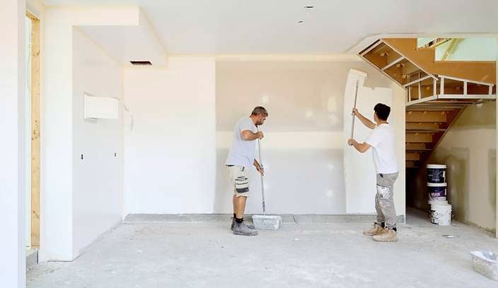 Two people painting interior walls of an unfinished room, using long-handled rollers. The space has bare concrete flooring, partially painted white walls, exposed sections around windows, and a wooden staircase with construction materials and paint buckets nearby.