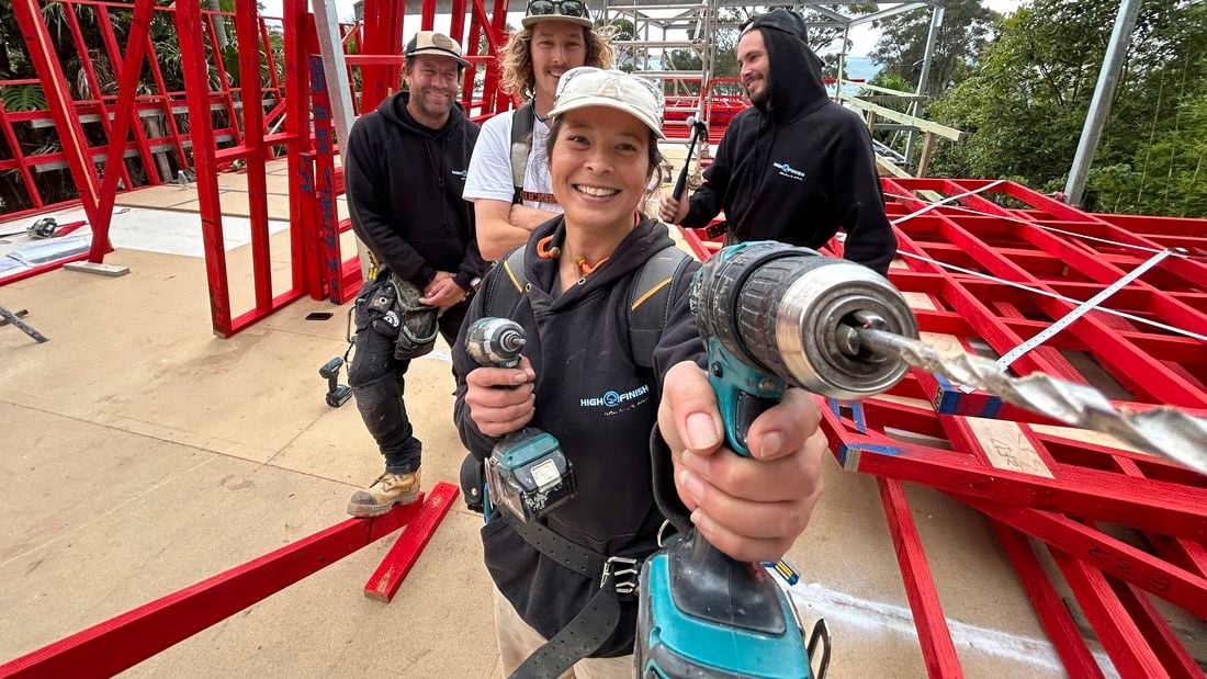 A female apprentice holds up a drill to the camera while three colleagues are behind her on a building site