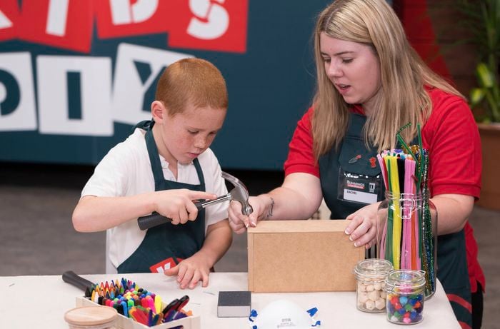 A Bunnings team member is helping a kid attach the front and back pieces of a tool caddy with a nail and hammer in a Bunnings DIY workshop.