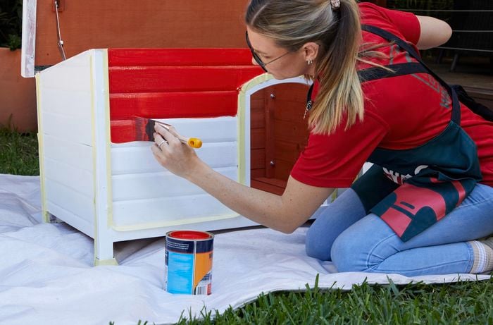 A woman paints a dog kennel red using a paint brush and exterior paint