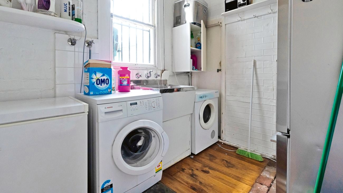 Old laundry with bland colour scheme featuring washing machines, dryers and shelves full of detergent and other cleaning products.