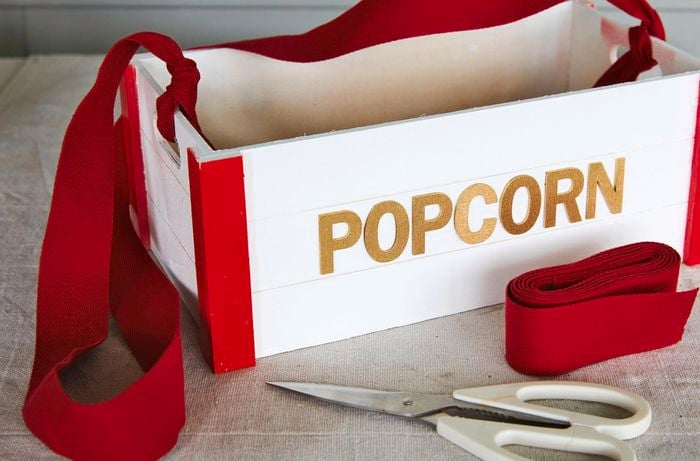 A craft wooden box labelled ‘popcorn’ with the corners painted red and straps attached to the handles. Scissors and red ribbon sit in front of it.