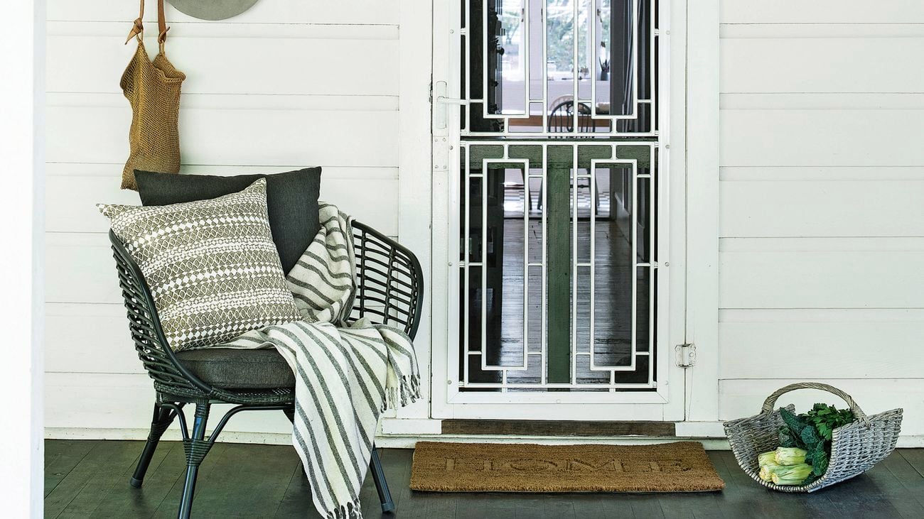 White weatherboard porch with a white metal security door and dark timber floor; green leaves hang down at the top. A woven chair with cushions and a striped throw sits beside wall hooks with a hat and bag, a doormat in front, and a basket on the floor.