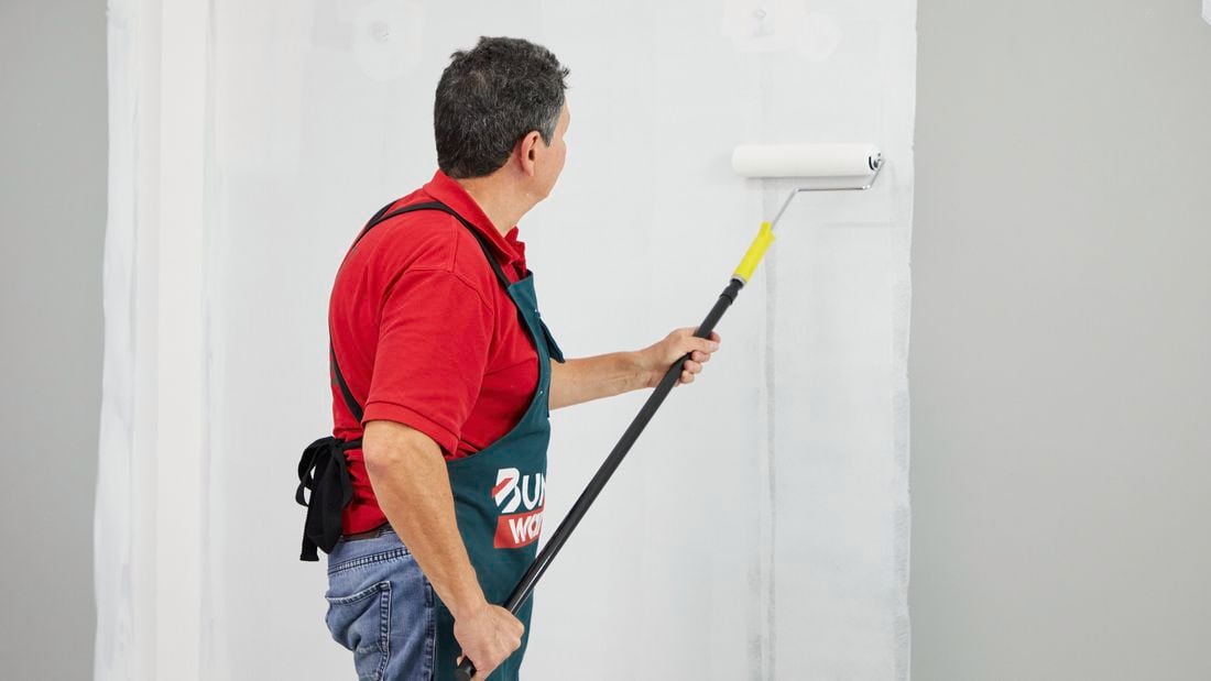 A man uses a paint roller to apply an undercoat to an interior wall.