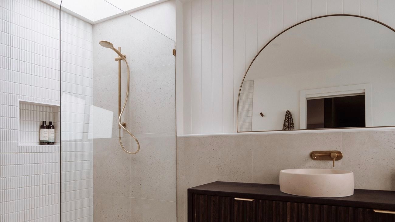 Modern bathroom with white shower, gold fixtures, a dark brown cabinet with a light pink sink and a semicircular mirror.