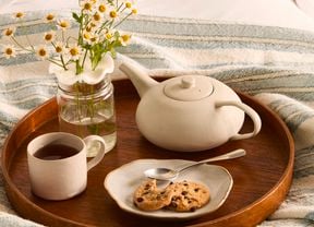 A wooden tray holding a glass vase with a flower frog holding chamomile flowers, a white mug of tea, a teapot and a plate with biscuits.