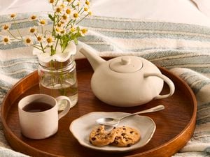 A wooden tray holding a glass vase with a flower frog holding chamomile flowers, a white mug of tea, a teapot and a plate with biscuits.