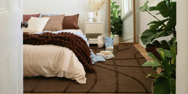Styled bedroom featuring soft neutral bedding, chunky throw, textured brown rug, indoor plants, and decorative mirror creating a warm, contemporary space.