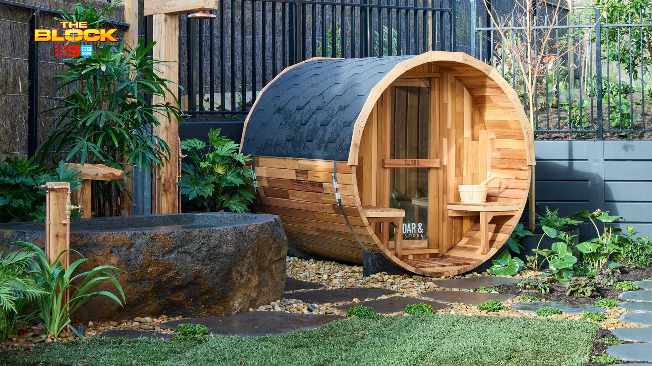Outdoor garden featuring a timber barrel sauna, rustic stone water trough, surrounding greenery and landscaped garden beds.