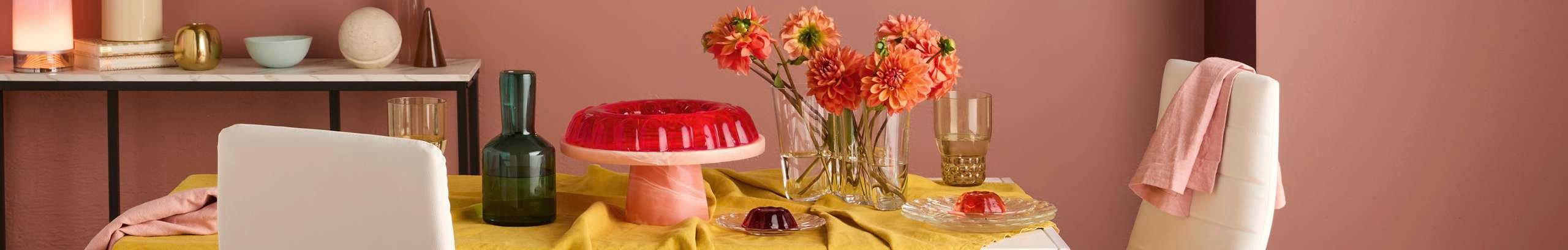 Styled dining table covered with a yellow tablecloth, featuring glassware, a green bottle, a pink cake stand with a jelly dessert, smaller jelly servings on plates, and a vase of bright orange flowers. Decorative objects and soft lighting sit on a console table against the wall behind.