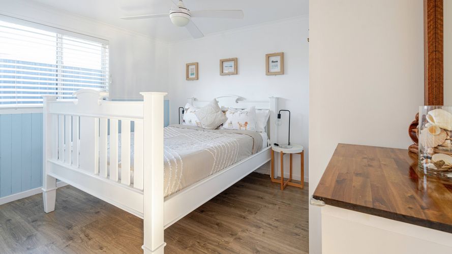 Bright coastal-style bedroom featuring a white four-poster bed, timber flooring, bedside tables and a large window with plantation shutters.