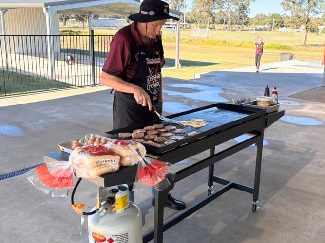 A man holding tongs standing behind a Barbecue with sausages and eggs on it.