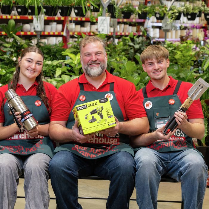 Three Bunnings team members seated among indoor plants in a store garden centre. One person holds a Ryobi ONE+ power tool kit, another holds a stainless-steel Thermos flask, and the third holds a barbecue tool set. Shelves of potted plants and hanging baskets fill the background.