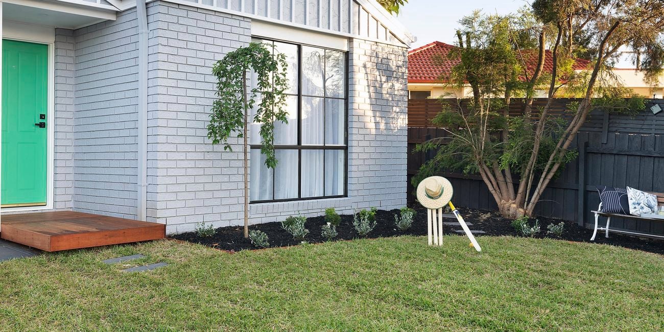 Front of brick house painted grey, with green lawn, neat garden and cricket set.