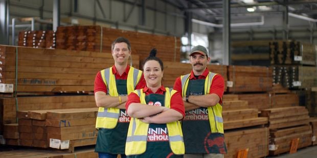 Group picture taken in the middle of a timber yard at a Bunnings store of three Bunnings team members who are our Landscape Specialists each dressed in Bunnings red uniform, green apron and hi-vis vest standing in a triangle formation crossing their hands together across their chest with each smiling at the camera.
