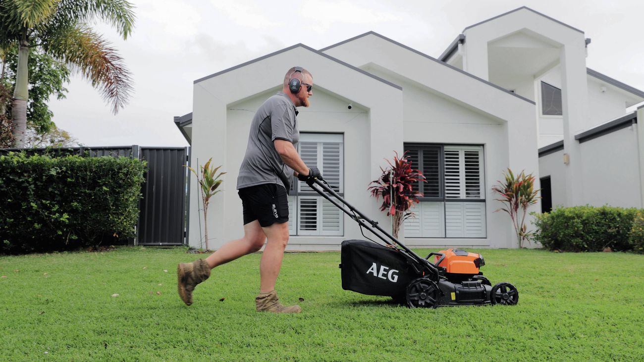Person mowing a residential lawn with a powered lawn mower beside a modern house.