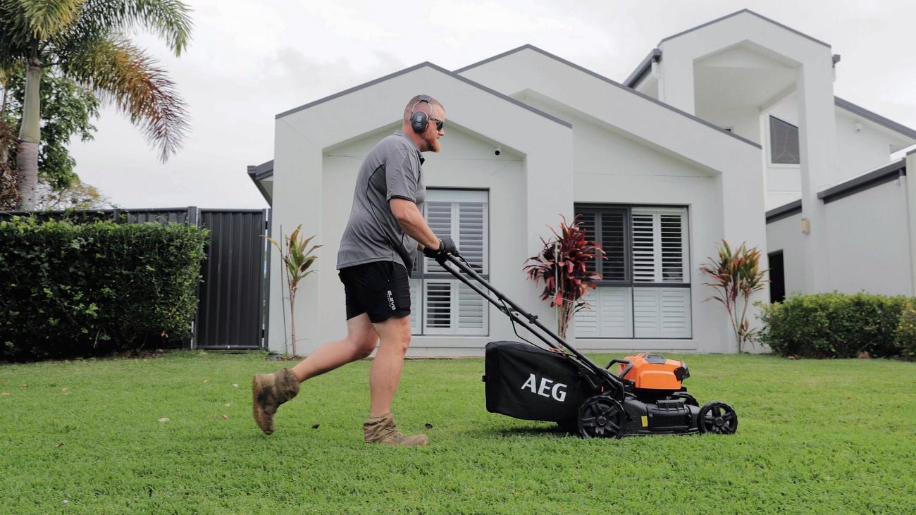 Person mowing a residential lawn with a powered lawn mower beside a modern house.