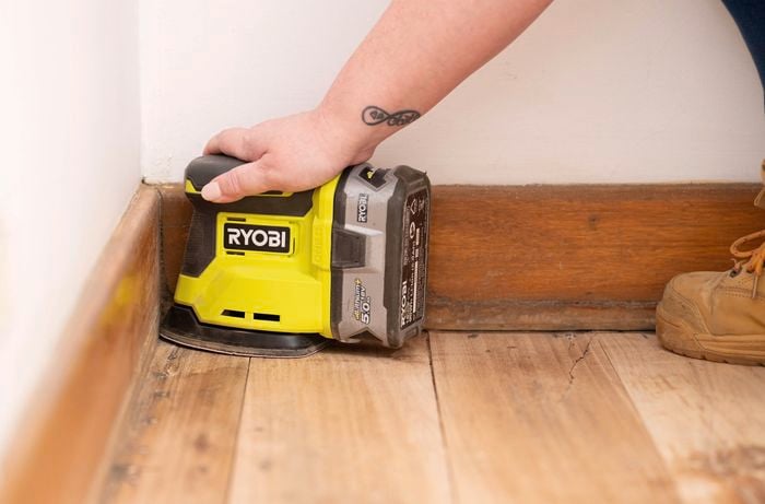 A person is using a palm sander ​to smooth timber floor corners near the skirting board to create a smooth and even finish​.