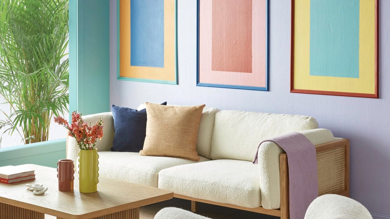 Living room with a cream colour sofa, three colourful rectangular painted wall panels on a purple wall, a tan coffee table and a cream colour bean shaped sofa chair