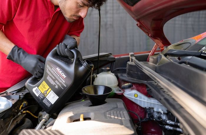 The image shows a man in a red shirt and black gloves carefully adding engine oil into a car engine using a funnel.