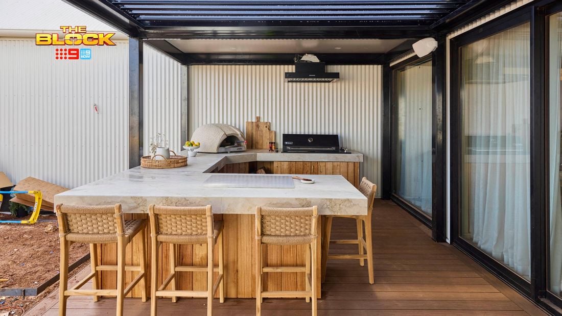 An outdoor area with a kitchen bench, with a pizza oven, barbecue on it and chairs in front of it.