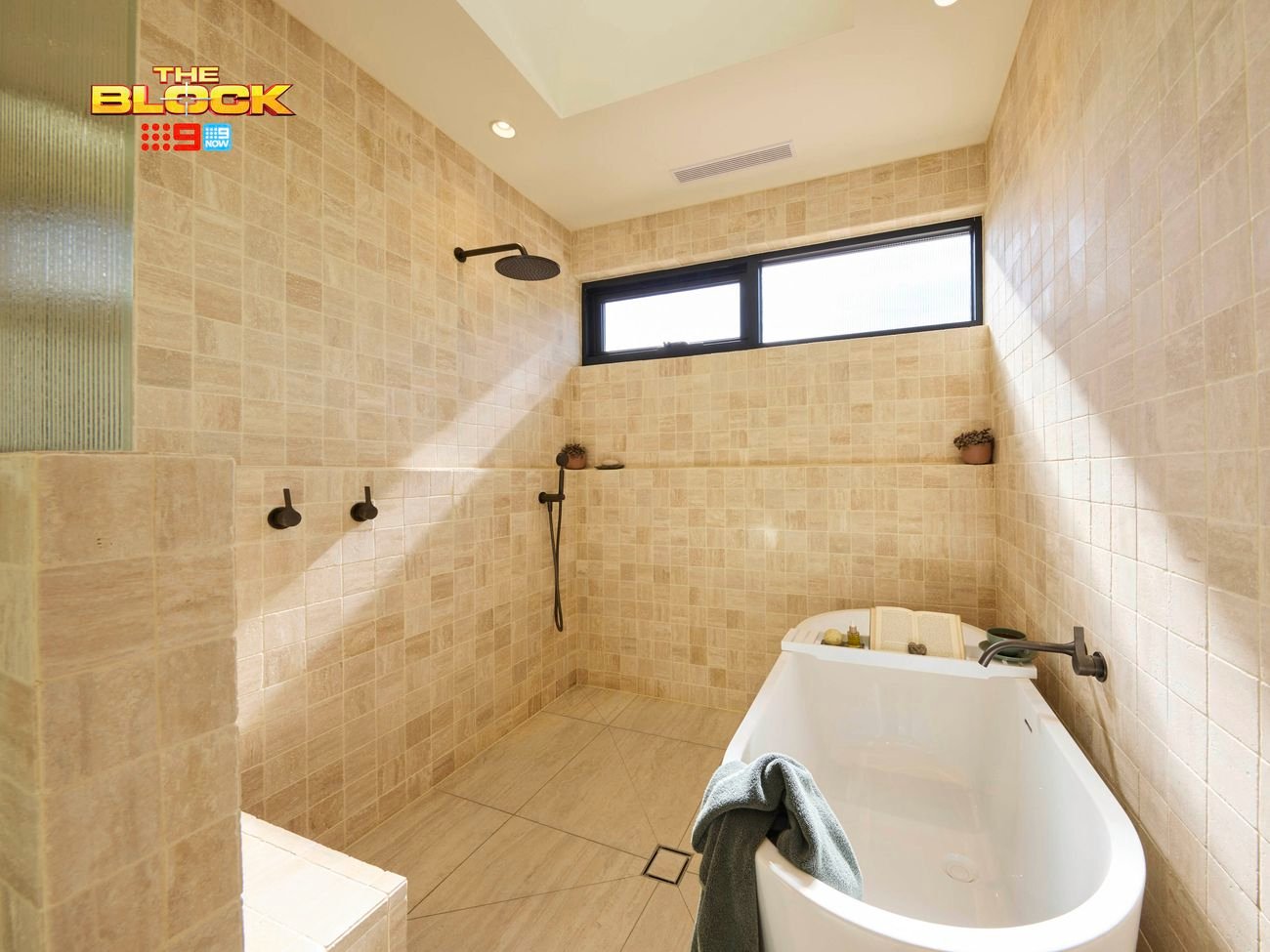 Modern bathroom with beige tiles, white bathtub, black showerhead and knobs, and natural light from a small window. 