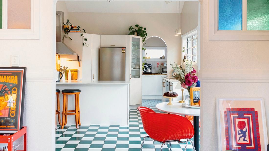 A vibrant kitchen with a green-and-white checkered floor, white cabinetry, a red dining chair and colourful stained-glass windows.