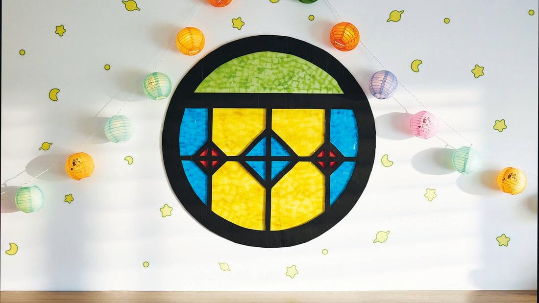 A round stained glass-look window made from black flute board with coloured cellophane as the panes, attached to a wall with paper lanterns and glow in the dark stickers