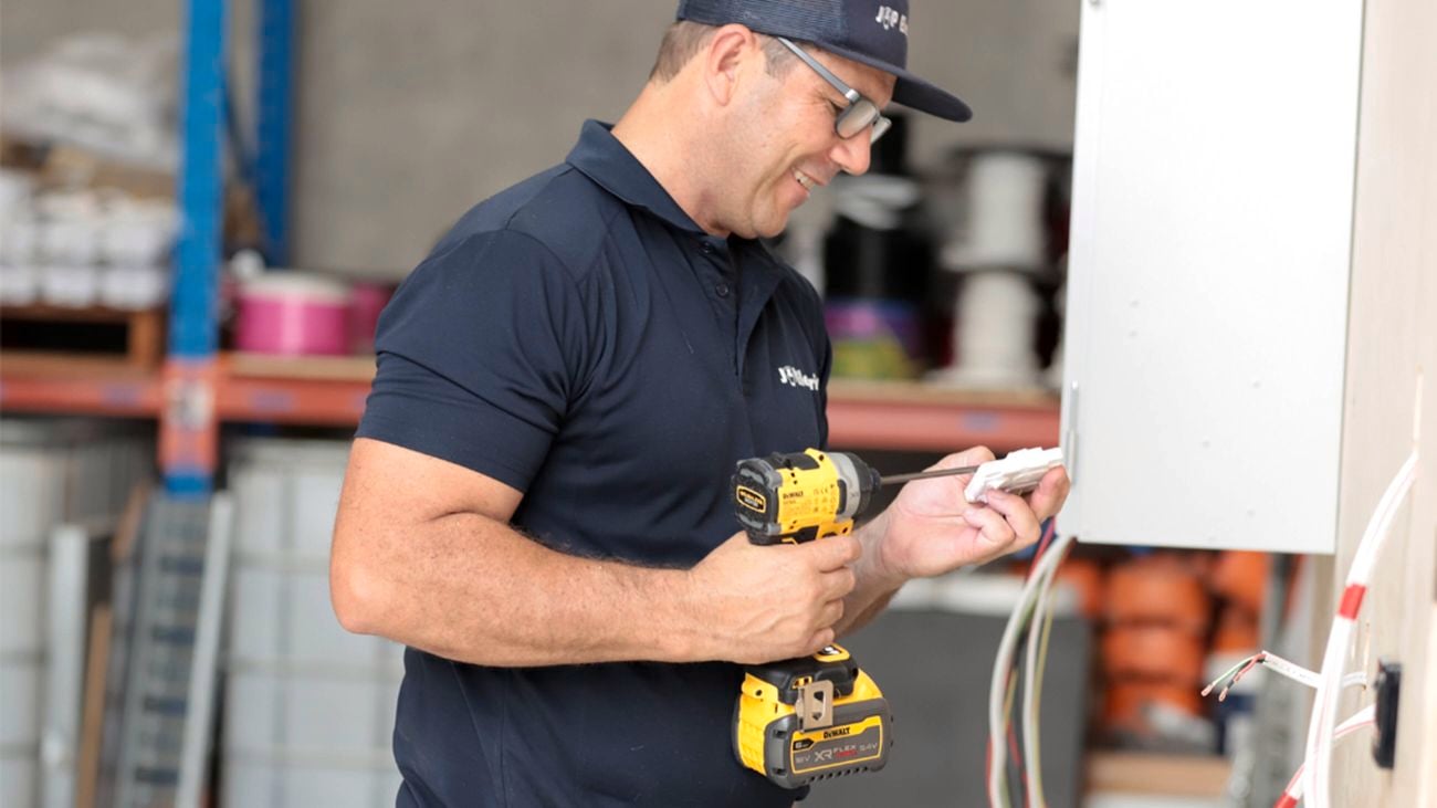 A close-up image of a man wearing a navy blue polo shirt and glasses uses a DeWALT drill to do some unfinished wiring work