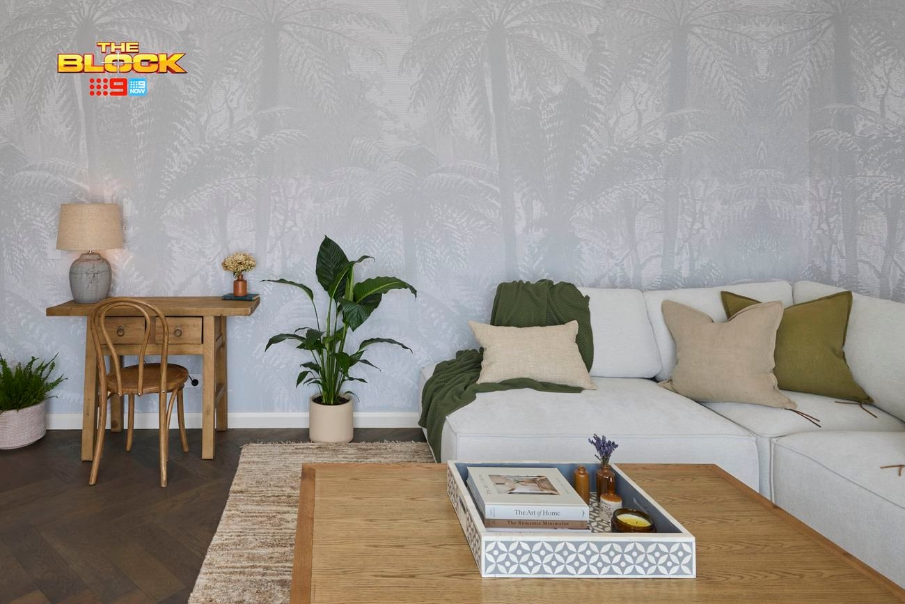 A well-decorated living room with light gray palm tree-patterned wallpaper. A wooden desk with a chair, table lamp, and decor sits on the left, next to a large green plant. On the right, an L-shaped white sofa with cushions and a green throw blanket faces a wooden coffee table holding books and decorative items. 