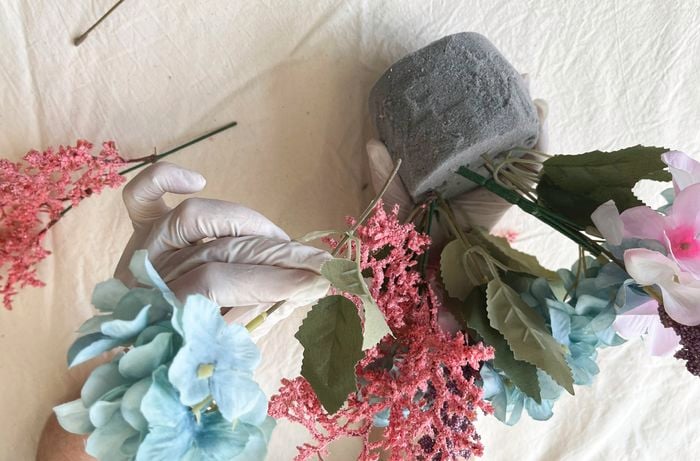 Hands wearing gloves apply craft glue to a small cut-out shaped like a bunny foot beside a floral arrangement.